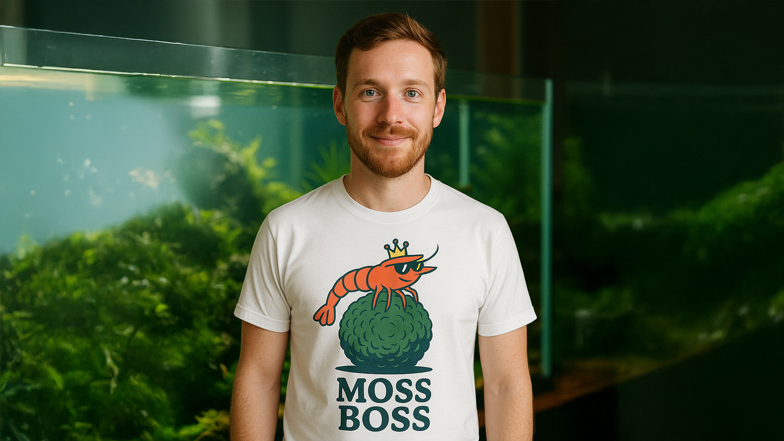 Lächelnde Person mit weißem Shrimplab-Moss-Boss-T-Shirt vor Aquascaping-Aquarium, freundlicher Blick zur Kamera.